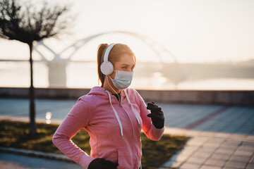 Fototapeta premium Beautiful young and fit woman in good shape running and jogging alone on city bridge street. She wearing protective face mask to protect herself from virus or allergy infection. Sunset in background.