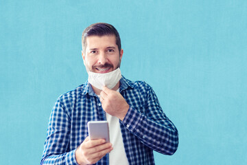 Confident mature man with big smile and open protective face mask using smartphone against wall