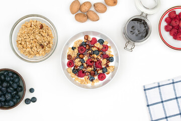 Natural yogurt with granola, fresh raspberries, blueberries and nuts with date syrup in a ceramic bowl on a white table. Top view on healthy breakfast or dessert with ingredients. 
