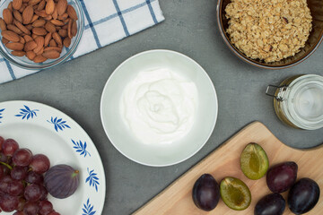 Natural yogurt in a ceramic bowl with plums, grapes, almonds, granola, agave syrup around. Top view on healthy breakfast ingredients on grey wooden table. 