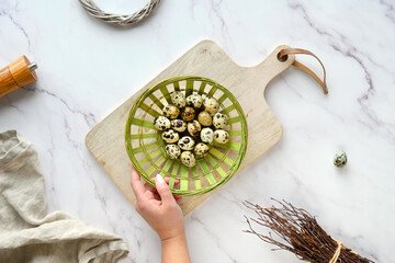 Hand holds rattan plate, basket with quail eggs. Flat lay on white marble table.