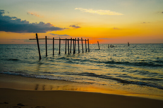 The view of the old wooden bridge decayed in the middle of the ocean in the evening. The sun is setting, making the sky a golden yellow. The waves hit the shore with orange light. There is copy space.