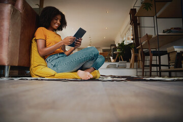 Low angle view of happy African American woman with afro sitting on floor with a pillow using a digital tablet