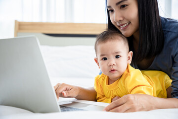 Young mother paly and searching data via laptop for baby daughter on the bed. Take care and help concept.