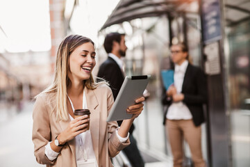 Young happy businesswoman holding digital tablet and coffee to go outside of modern building, businesspeople in background.