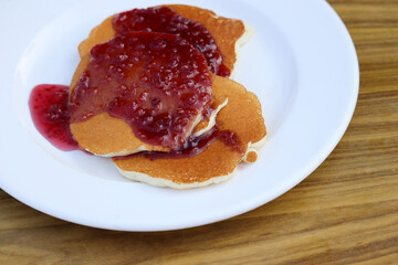 Pancakes drizzled with raspberry jam in a white plate on wooden table. Tasty dessert, sweet breakfast