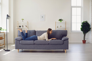 Fototapeta premium Young smiling woman lying on sofa at home and working online on laptop