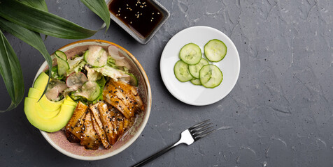 Hawaiian dish. Poke salad with eel, rice and avocado in a deep bowl on a gray background. Next to it is a white plate with slices of fresh cucumber. The view from the top. No people. Copy space