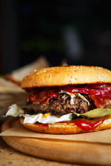 Big fat burger with bacon stripes served on wooden plate on table in American fast food restaurant.Delicious hamburger with pork meat strips & beef patty cooked on grill pan in diner cafe