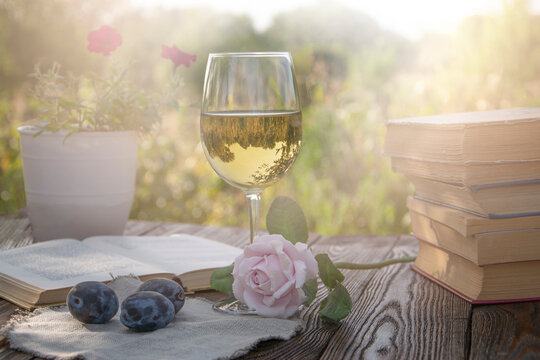 The photo shows a glass of white wine with a rose, plums and books, in the background of nature with a sunny sunset.