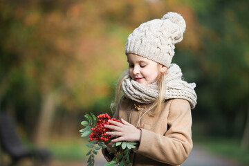 Cute little girl holds a branch of rowan tree with red berries in her hands and touches  it. Pretty blond girl wearing beige coat, knitted cap  and scarf explore the world in autumn park, close-up.