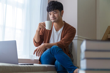 Portrait of an attractive smiling young man wearing casual clothes sitting beside window at home, using laptop computer.