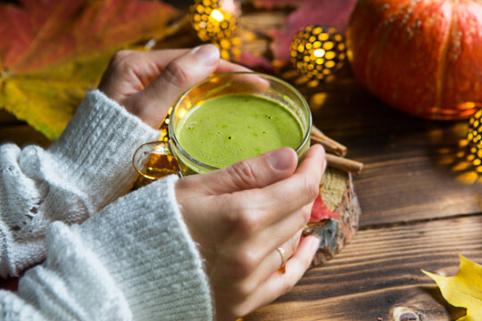 Green Japanese matcha tea with foam in transparent Cup on wooden table in autumn still life. Women's hand with long white sweater sleeves are warmed by drink. Focus on Cup. Warm atmosphere and comfort