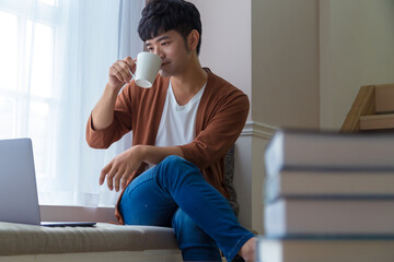 Portrait of an attractive smiling young man wearing casual clothes sitting beside window at home, using laptop computer.