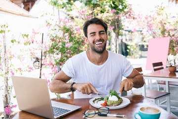 Half length portrait of cheerful caucasian male eating healthy meal satisfied with brunch laughing in good mood, positive funny hipster guy keeping healthy lifestyle enjoying vegan food in cafe