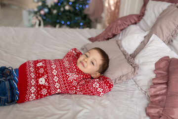 Obraz na płótnie Canvas Happy little boy in red knitted sweater, dream on about presents laying in the bed. Christmas tree background. Enjoy winter holidays.