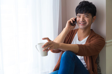 Handsome man is siting near window with cup of coffee and smart phone in hands. Talking on mobile phone. Working at home concept.
