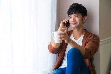 Handsome man is siting near window with cup of coffee and smart phone in hands. Talking on mobile phone. Working at home concept.