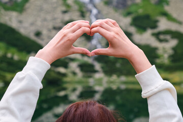 Obraz premium Close up woman hands showing a heart on the amazing background of waterfall and transparent, clean, water of the lake with copy space. Vocation in the mountains concept. Solitude with nature. 