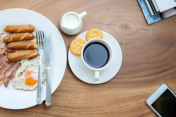 Top view of breakfast table with bacons, sausages, fried egg, coffee with milk and biscuits on white dish. The chili sauce and ketchup to enhance the flavor. Sprinkled with ground black pepper on top.