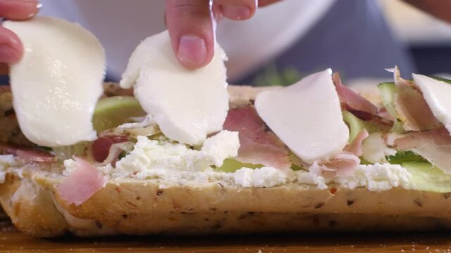 Young chef puts thin slices of mozzarella cheese on sandwich in kitchen on cutting Board in home interior. Preparation of morning Breakfast from bun cucumber cheese ham. Baguette sandwich.