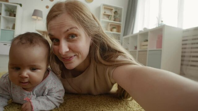 Close up of joyful Caucasian woman with long hair lying on carpet in living room and making selfie with her adorable mixed-race toddler lying nearby