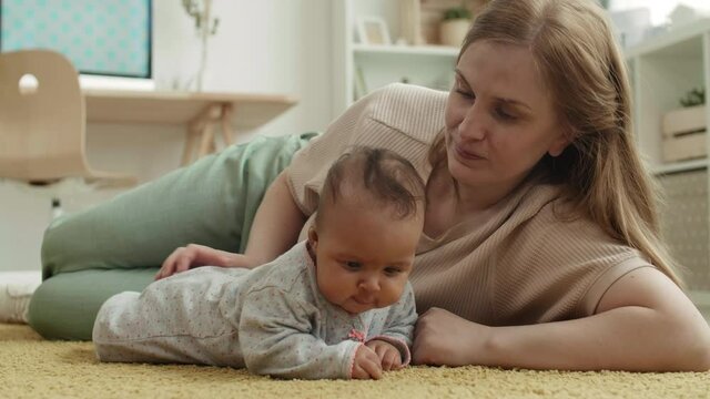 Lockdown of Caucasian woman with long hair and cute mixed-race baby girl lying together on carpet at home. Mother kissing her baby