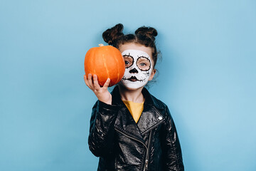 Headshot of beautiful little girl with skull makeup, holds pumpkin near face, wears black clothes, looking at camera, poses against pastel blue studio background with copy space. Happy Halloween!