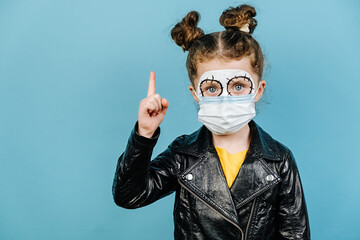 Little girl with bright scary makeup wears protective medical mask, points away, demonstrates something at blank copy space for your advertisment, isolated over on blue background. Happy Halloween!