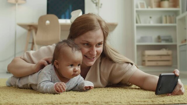 Lockdown of happy Caucasian woman with long hair lying on carpet at home and making selfie with her cute mixed-race baby girl lying nearby