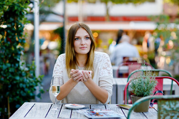 Fototapeta premium Young beautiful woman drinking cappuccino and cup of coffee and glass champagne while waiting for friend and breakfast. Happy alone woman in outdoor cafe or restaurant on sunny summer day.