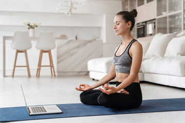 Fit young woman practicing meditation at home on exercise mat in her living room, repeating online...