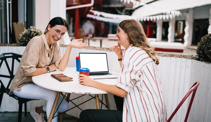 Cheerful female freelancers discussing mockup website from computer and grinning, joyful distance colleagues using blank netbook with copy space area during remote work in sidewalk with coffee to go