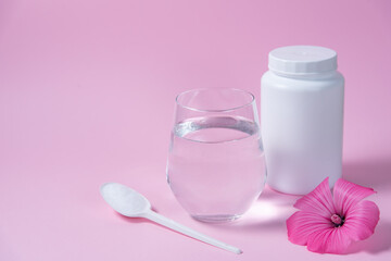 spoon of collagen powder from a white jar with a glass of water on a pink background