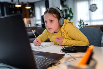 Cute child with headphones using laptop computer for online learning. Home school. Girl doing homework at home. Lifestyle concept for home schooling. Social distancing and education.