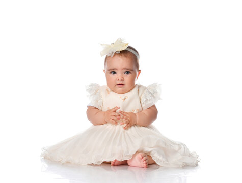 Beautiful baby in a festive dress and a hair band sitting on the floor. Toddler toddler looking at camera studio portrait shot on white background. Children's fashion and beauty