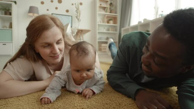 Close up of joyful Caucasian woman, African-American man lying on carpet in living room and making selfie with their adorable toddler lying nearby