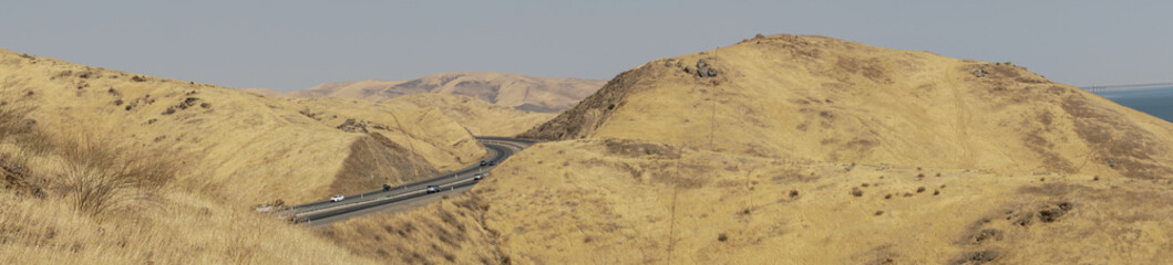 Panoramic view of the freeway road with cars crossing the the San Luis Reservoir valleys during dry season, San Luis Creek in the eastern slopes of the Diablo Range of Merced County, California. USA