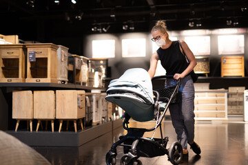 Fototapeta na wymiar Young mom with newborn in stroller shopping at retail furniture and home accessories store wearing protective medical face mask to prevent spreading of corona virus. New normal during covid epidemic.
