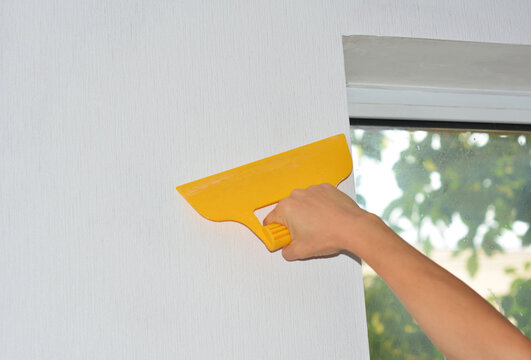 A professional wallpaper installer is wallpapering the wall around the window and removing air bubbles using a smoother tool.