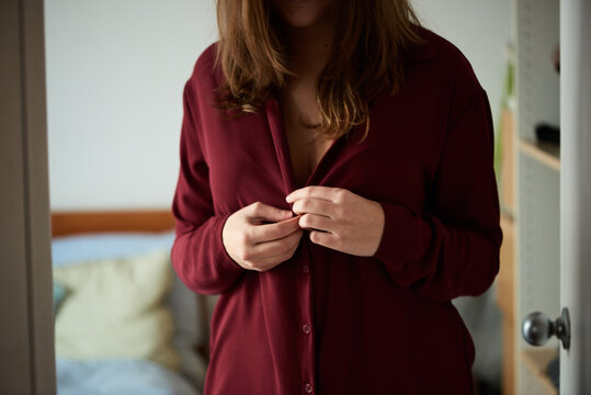 Brunette woman buttons red blouse up in bedroom.