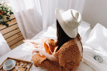 Beautiful young woman in white hat sitting on bed near window in sunny morning. Relaxing time