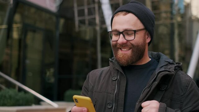 Crop View Of Man In Glasses Looking At Phone Screen And Smiling While Walking At Street. Bearded Guy With Bag On Shoulder Scrolling Social Media News Feed While Using Smartphone.