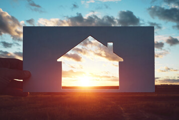 Man holds a piece of paper with a house cut out in it against the sunset.Home in nature, travel and housing. Concept.