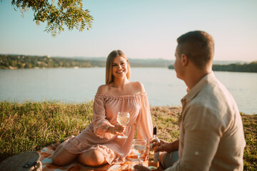 Fototapeta premium A cute blonde woman in a pastel pink dress and a caucasian man drinking white wine on the beach at sunset. A loving couple celebrating and enjoying a picnic on the river bank