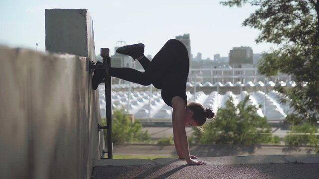Young female gymnast or yogi doing handstand with legs split outdoors. Slim flexible Caucasian woman training at the background of city at sunrise.