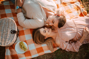 Top view of a caucasian man and a blonde woman  lying on an orange blanket at a picnic,  and next to them is a basket of food and grapes