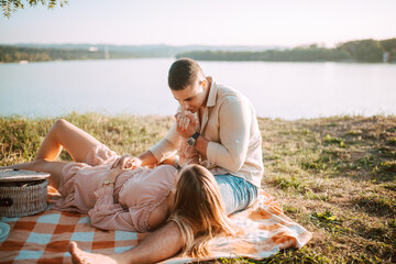 Fototapeta premium A beautiful blonde caucasian woman in a dress is lying on a blanket next to her man. Romantic loving couple on a picnic by the river