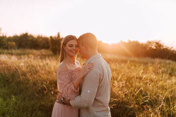 A beautiful blonde caucasian woman in a pastel pink dress and a caucasian man stand in a field at sunset.A loving couple in the nature