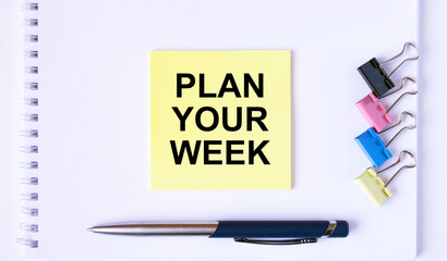 Yellow sticker with text Plan Your Week lying on a white Notepad with a pen and paper clips